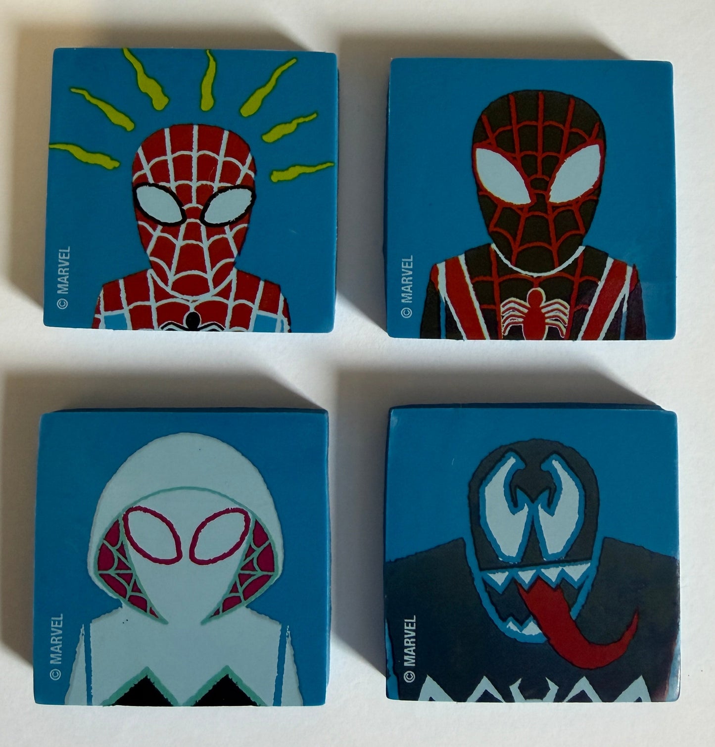 Four small square blue erasers featuring stylized Spider-Man illustrations, arranged in a 2x2 grid on a plain white background.